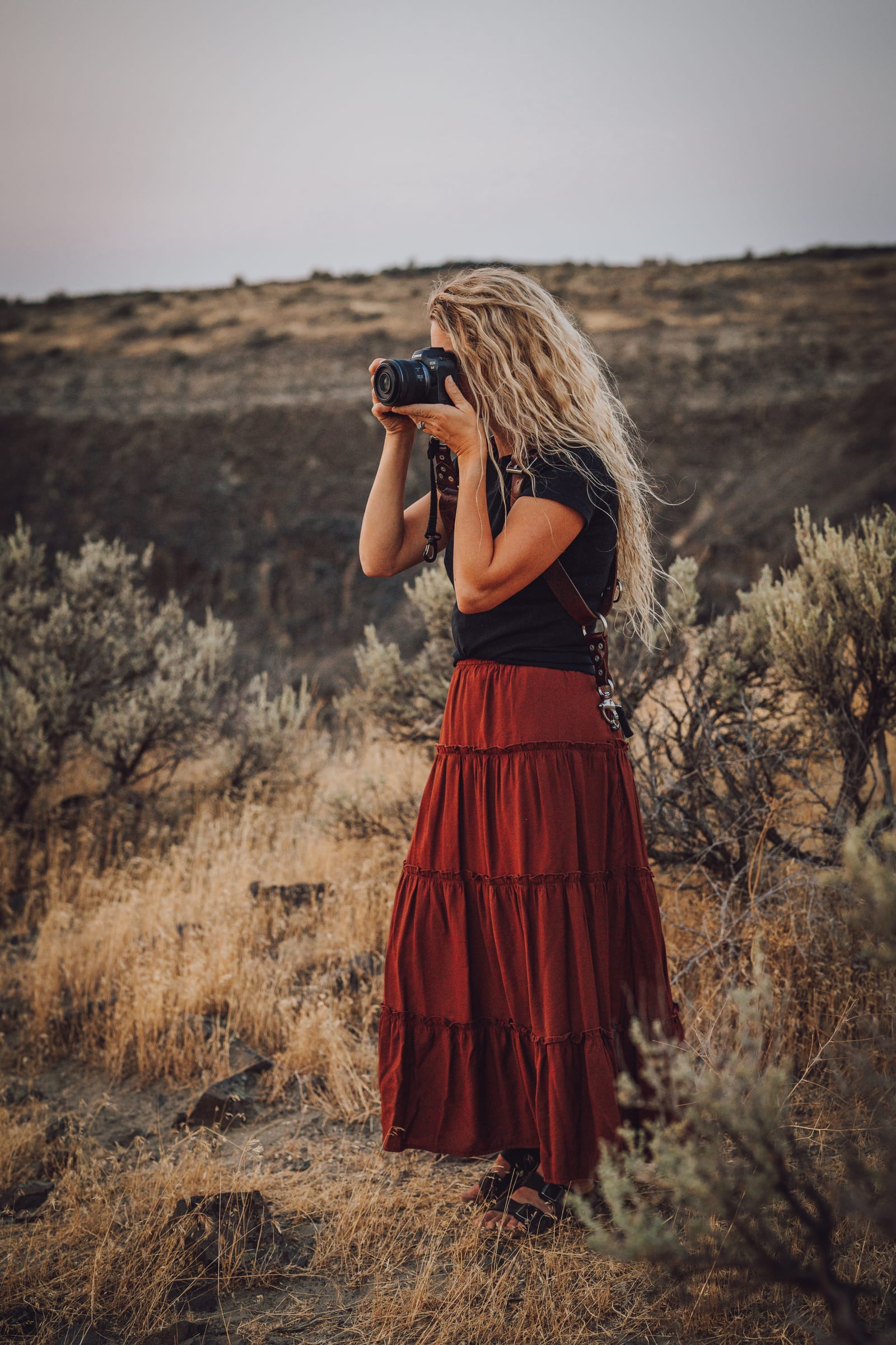 Lisa holding a camera to her face among dry grass and shrubs.