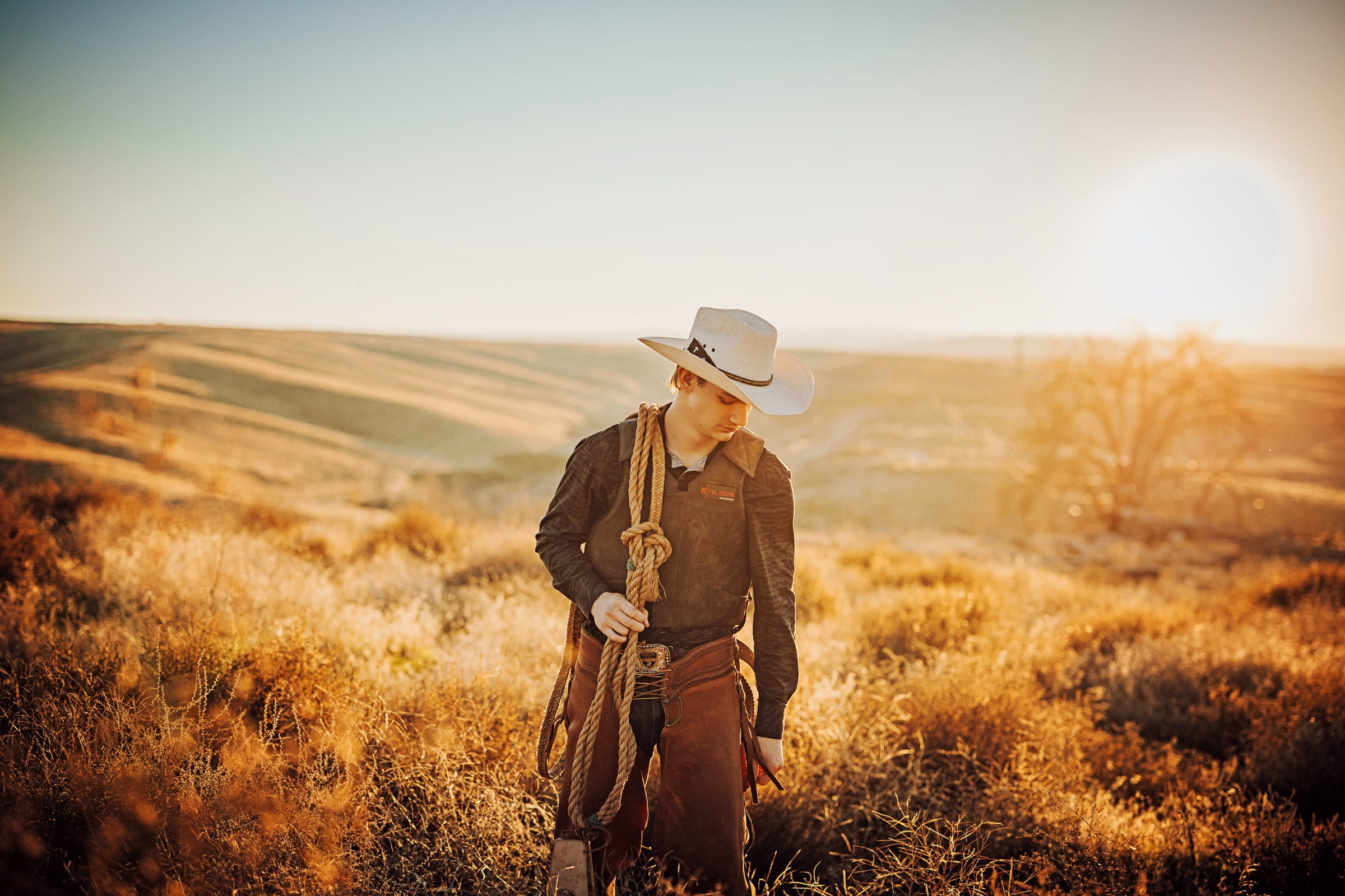 High school senior in a cowboy hat holding a rope in a golden field.