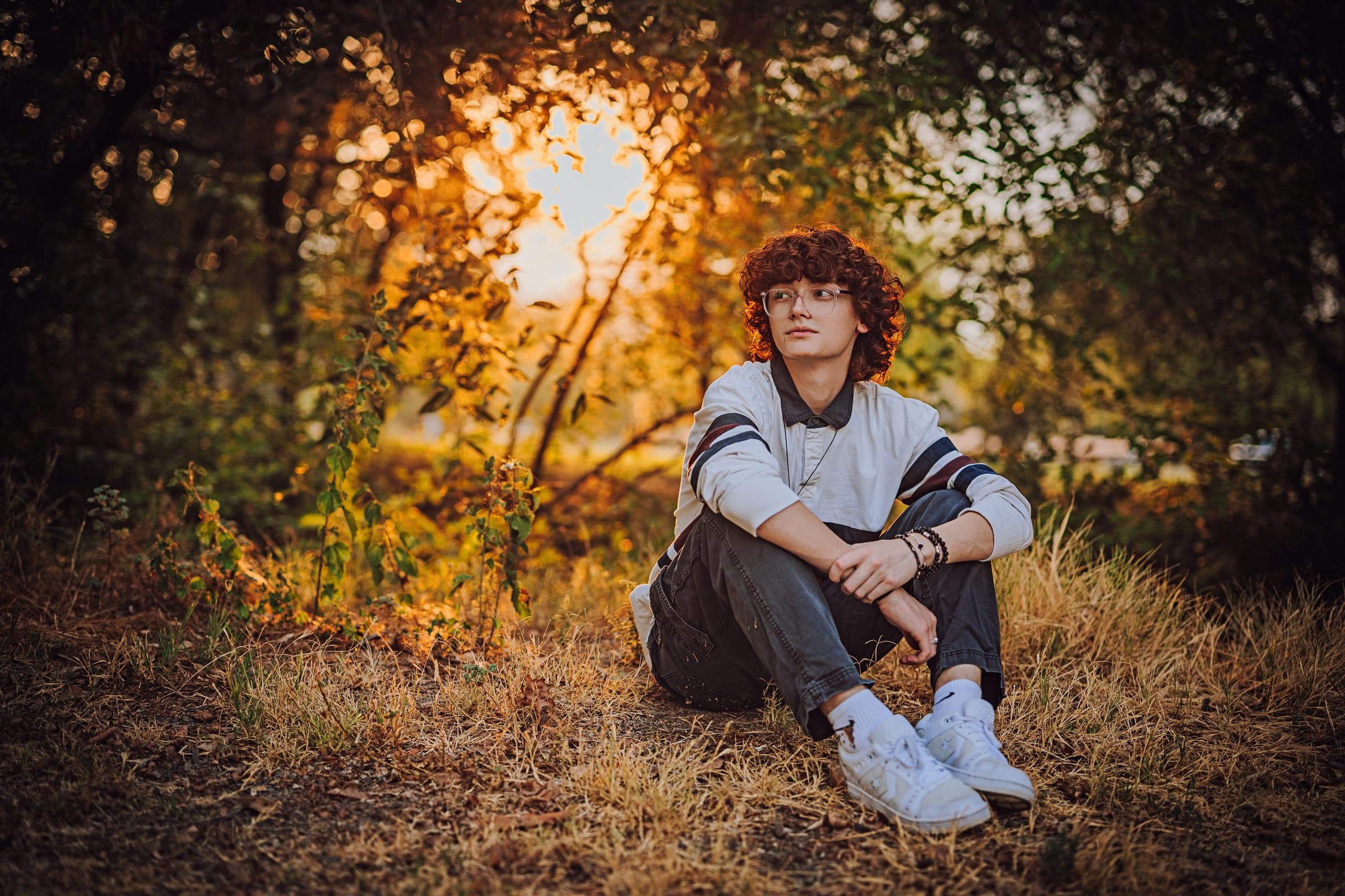 High school senior with glasses seated in dry grass beneath autumn trees.