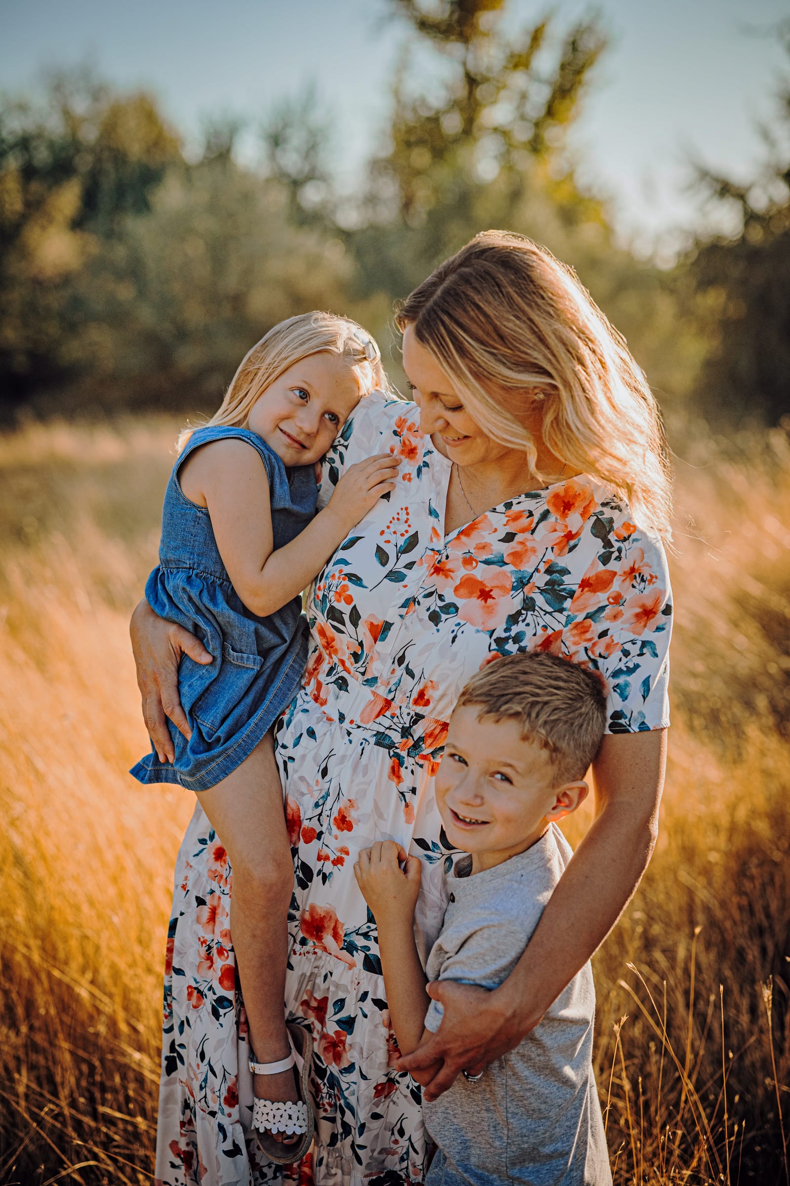 A woman holding a young girl while a boy stands beside them in warm evening light.