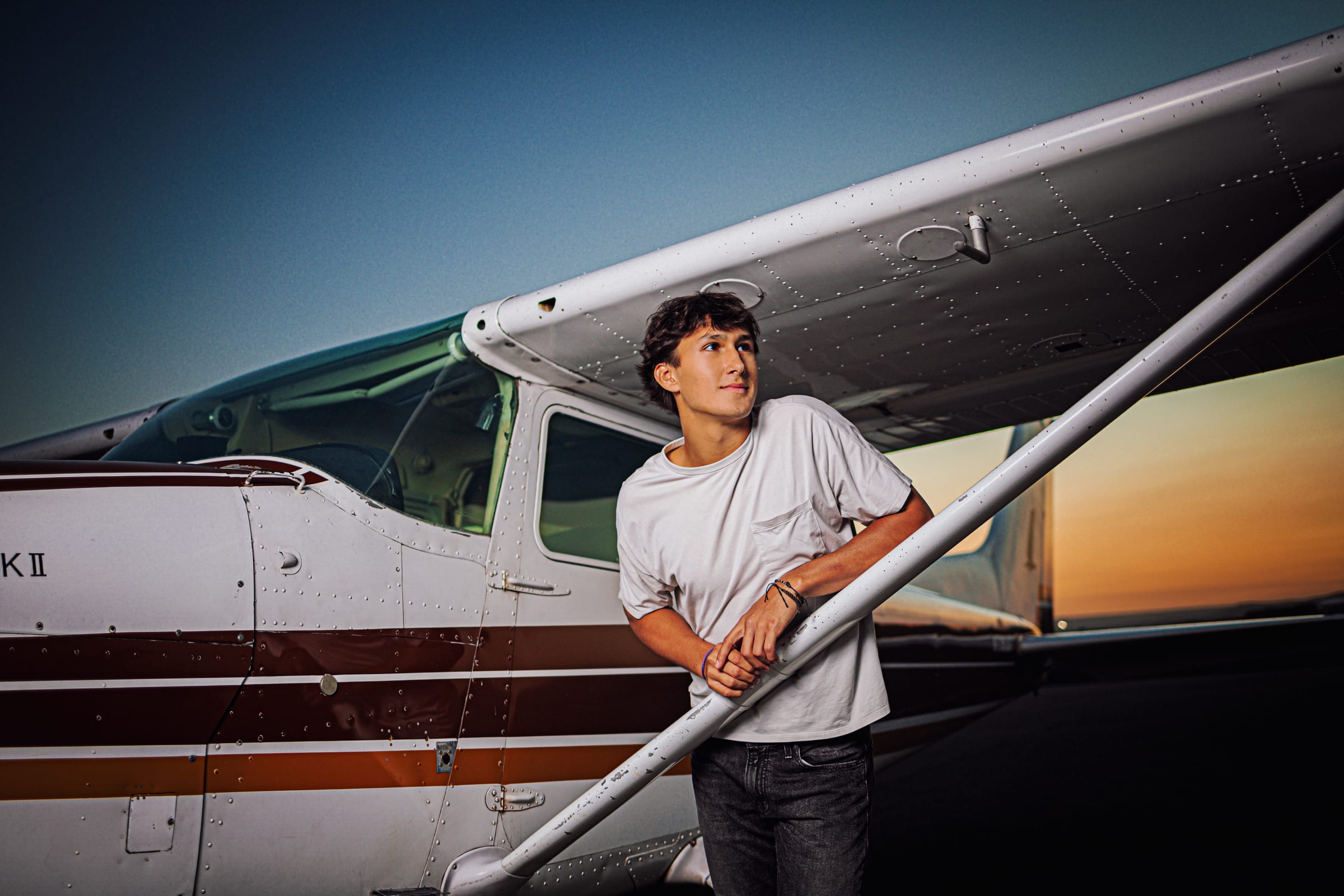 High school senior in a white shirt leaning beside a small airplane.