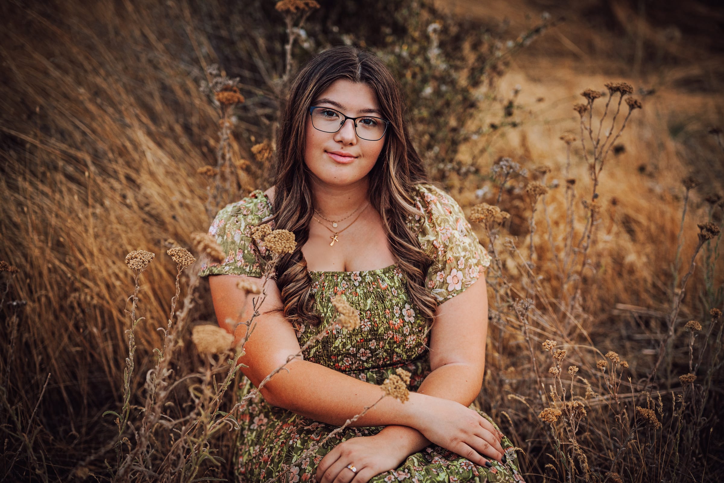 High school senior with glasses seated in dry grass wearing a green floral dress.