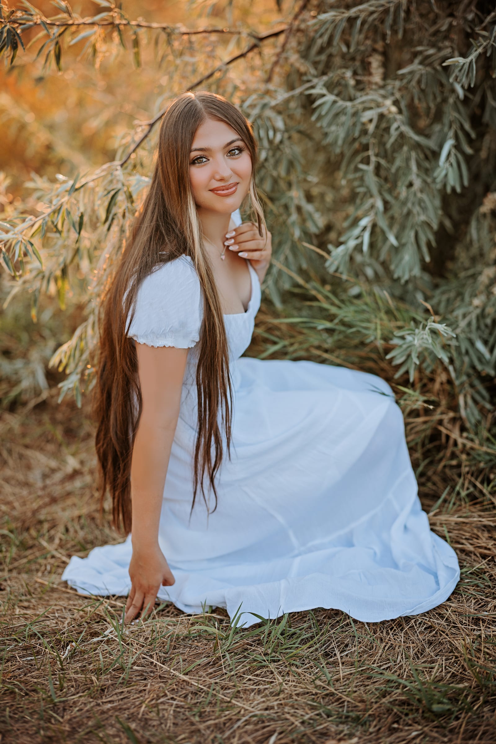 High school senior in a white dress seated beneath leafy branches.