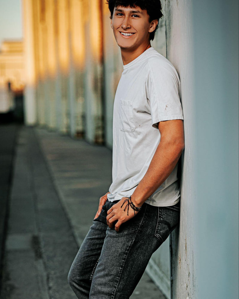 A young man in a white T-shirt leaning against an industrial wall