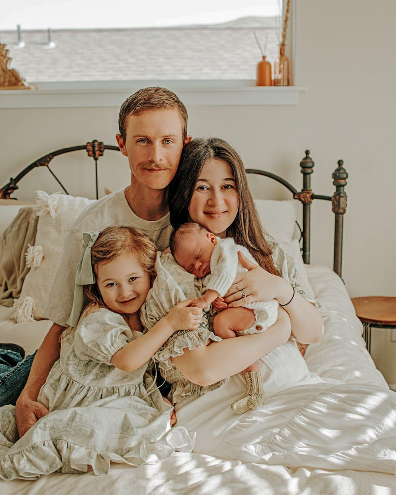 A family of four holding a sleeping newborn together on a bed