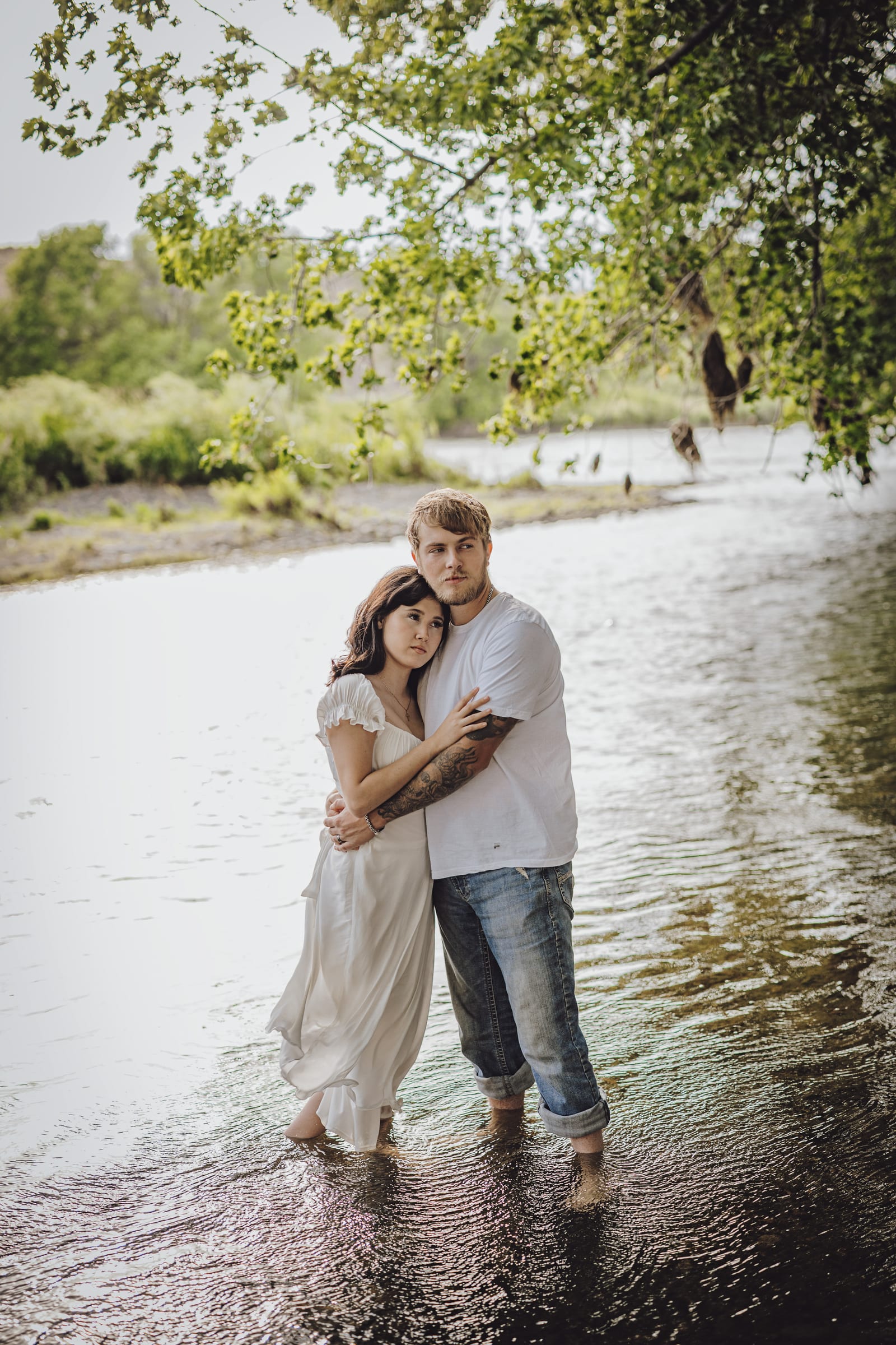 Couple standing together in shallow river water beneath leafy trees.