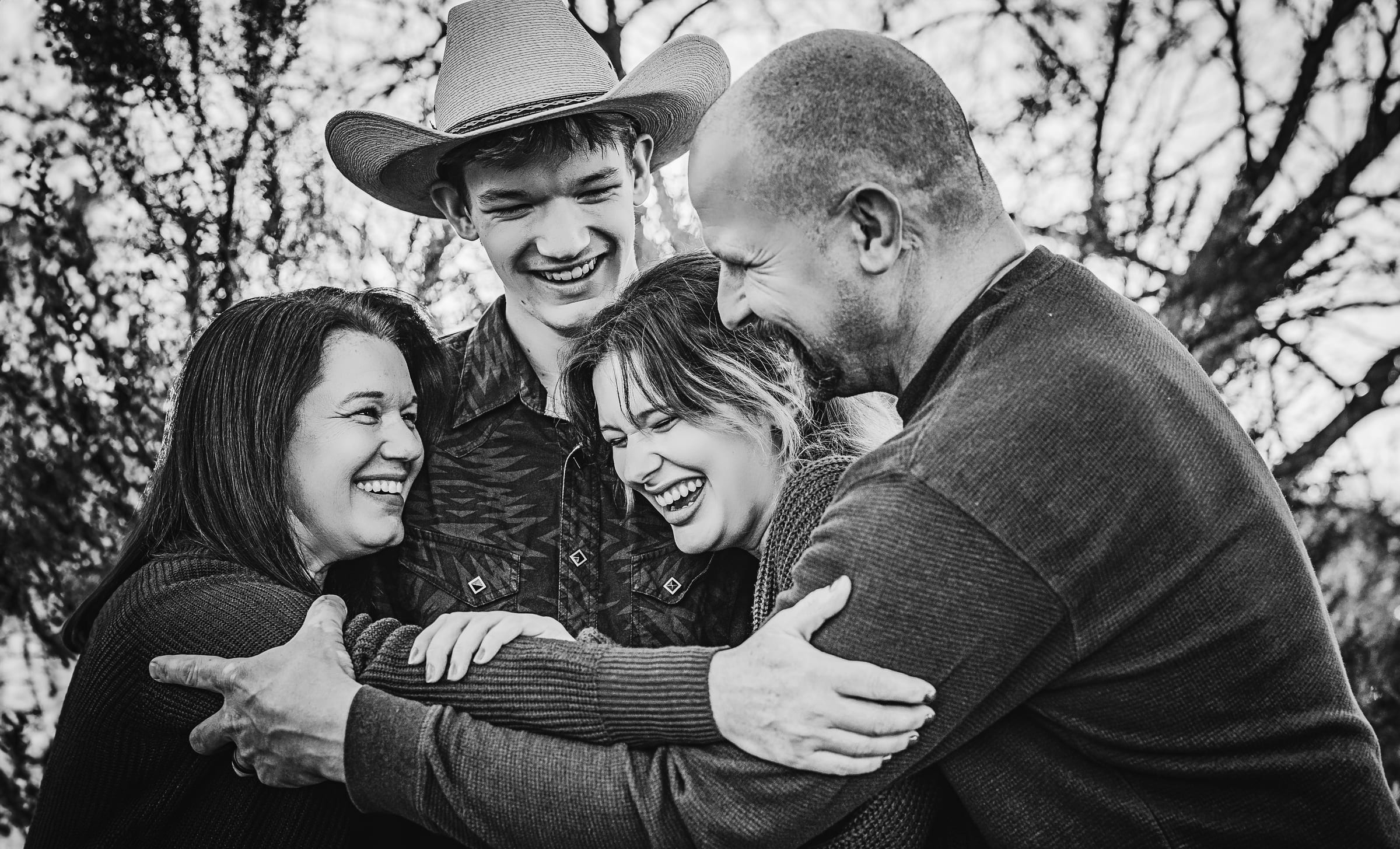 Family laughing together beneath bare trees in a black-and-white portrait.