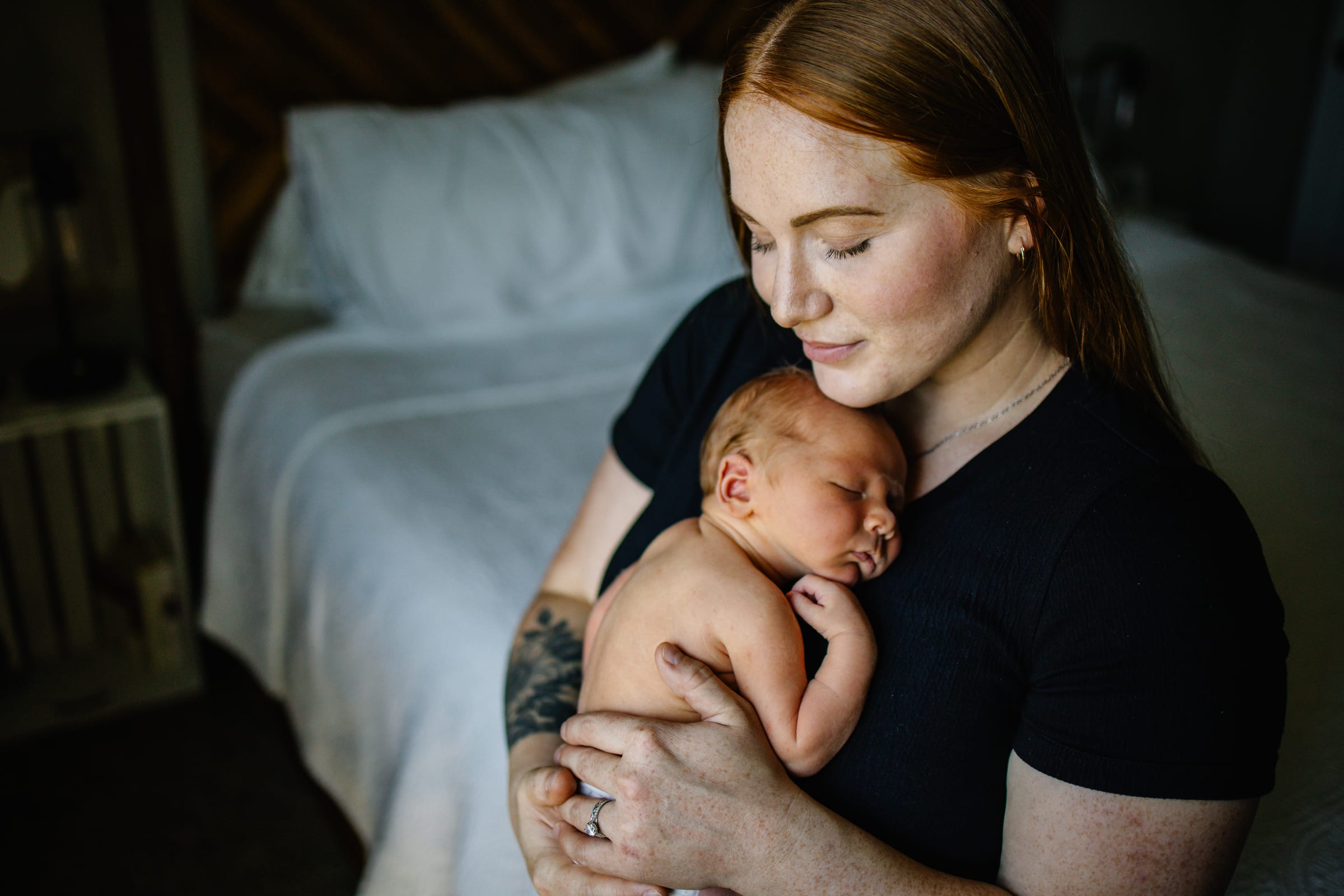 Mother holding a sleeping newborn beside a bed.