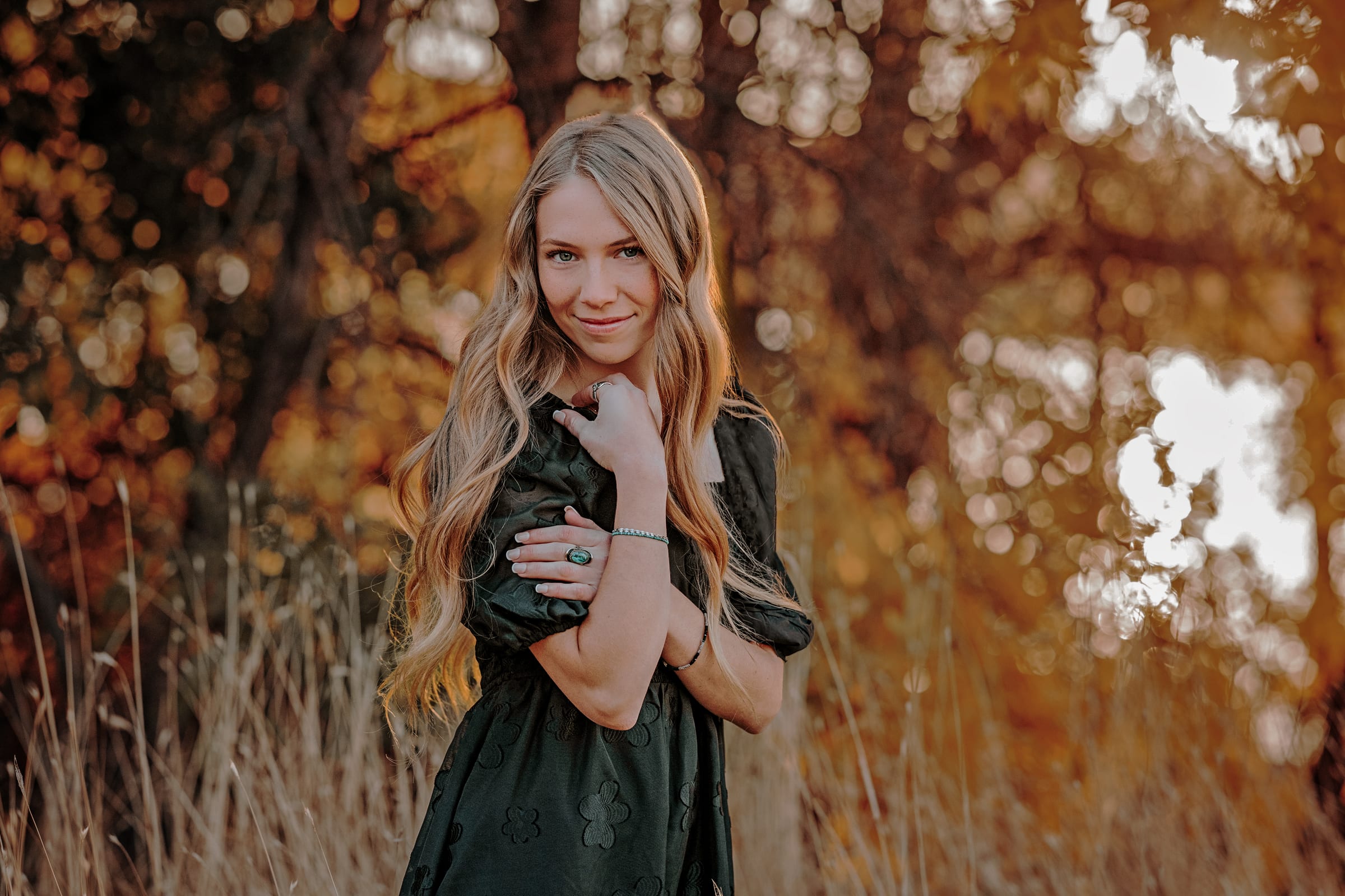 High school senior in a dark dress standing among autumn leaves.
