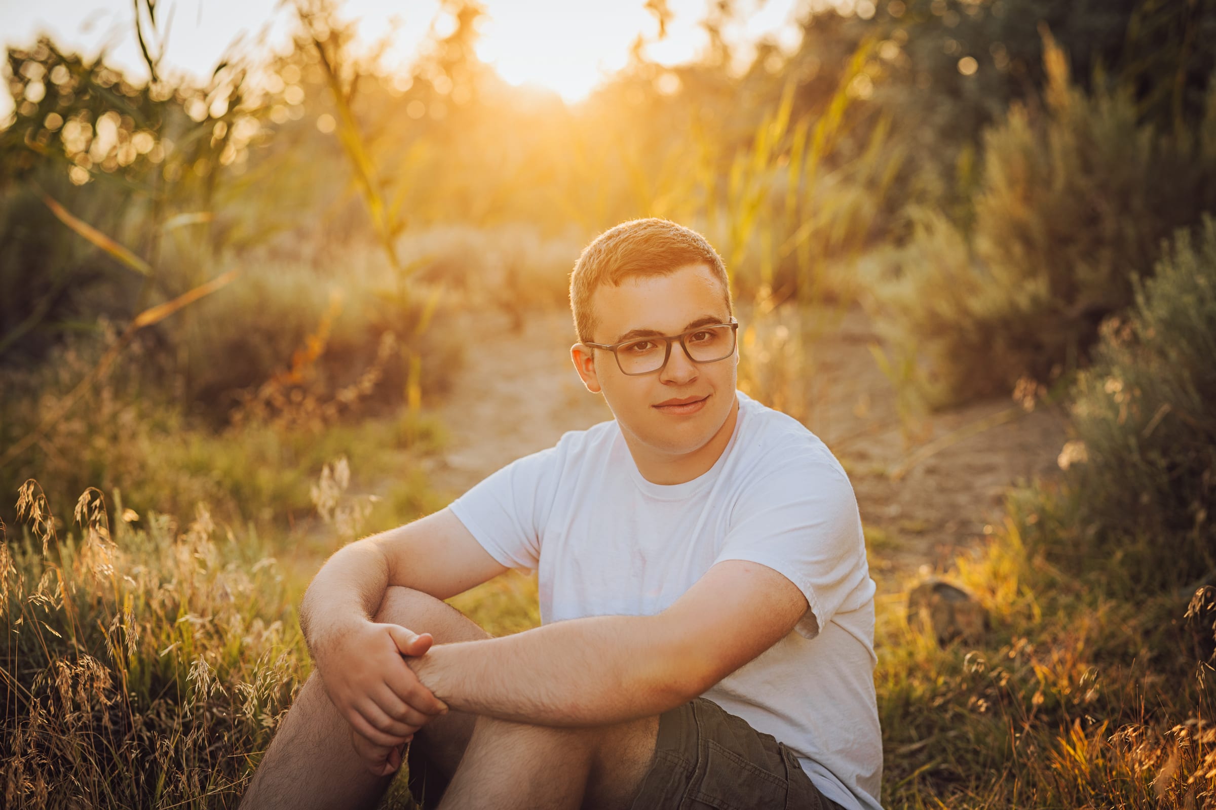 High school senior with glasses seated in warm evening grass.