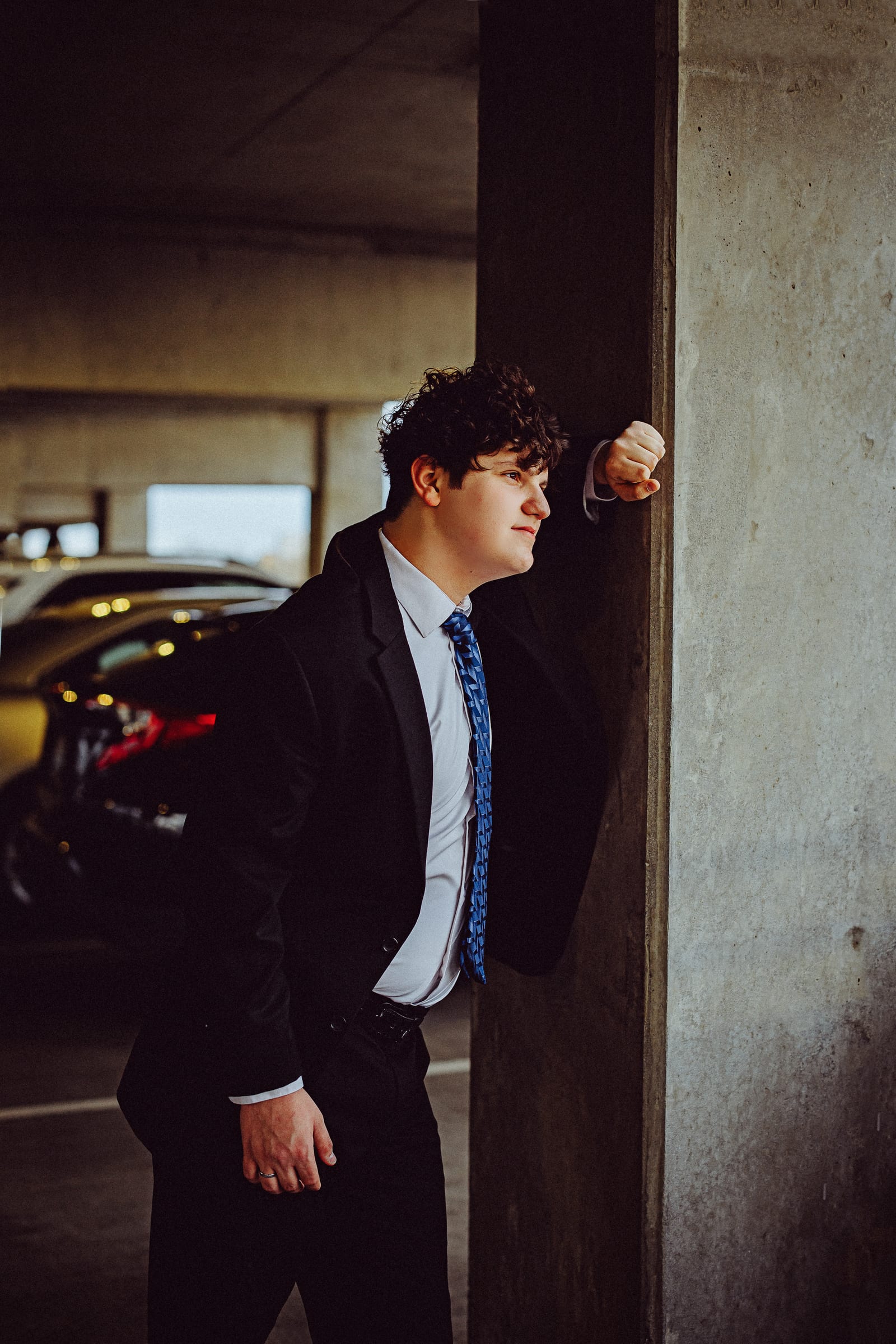 High school senior in a dark suit leaning against a concrete column.